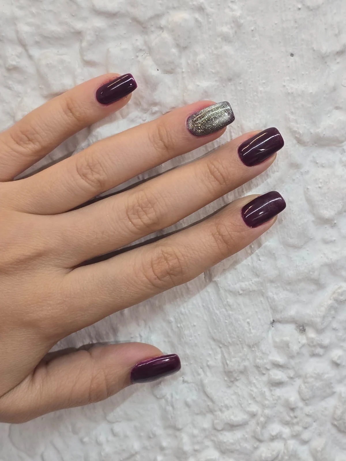 Deep plum glossy square nails with gold-glitter accent nail against white render wall