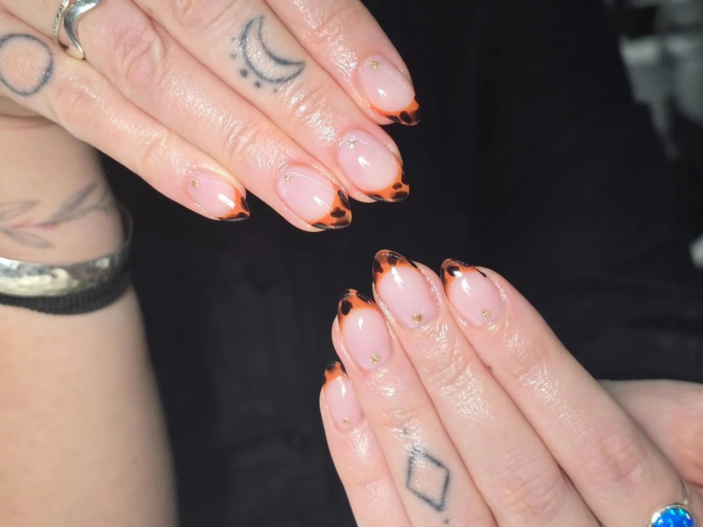 Tortoiseshell French tips on nude almond nails with gold-dot accents and tattooed fingers