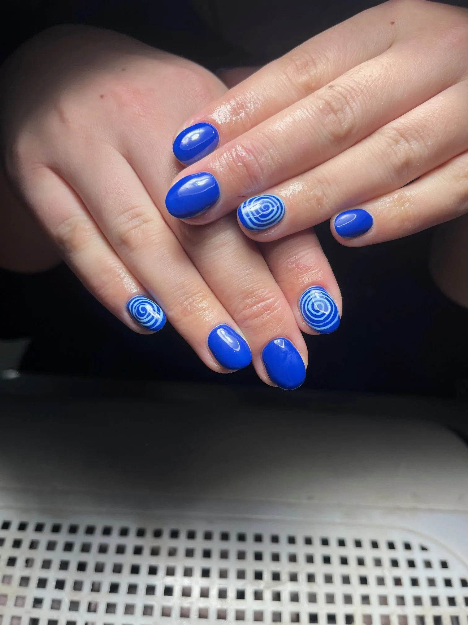Glossy cobalt-blue nails with white spiral art under salon light